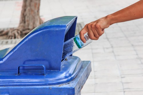 A person's hand is seen disposing of a plastic bottle into a blue recycling bin, which is situated outdoors on a paved surface with a tree trunk visible in the background. The recycling bin has a hinged lid open to reveal the top of the container, and it appears to be made of durable plastic with a textured finish. The person's arm is extended, and they are holding the bottle by the neck, preparing to drop it into the bin. The environment suggests a public or communal area, possibly in a residential or commercial setting in Willesden, where waste collection services like those from Commercial Waste Willesden are often utilized for rubbish disposal and recycling management. The scene highlights responsible waste sorting and recycling practices common in urban or suburban waste management contexts in northwest London.