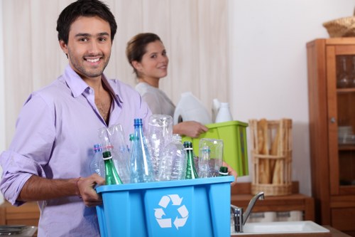 A young man with dark hair, wearing a light purple shirt, is standing in a domestic kitchen holding a blue recycling bin filled with empty glass bottles and jars, some clear and some green, with the recycling symbol printed on the front. Behind him, a woman with brown hair tied back, dressed in a light-colored top, is partially visible and appears to be standing near a green box used for sorting recyclable waste. The kitchen background includes a wooden cupboard on the right, and a countertop with a stainless steel sink beneath a window, illuminated by natural light. The scene suggests a household recycling activity, with the focus on the bin's contents and the domestic interior environment, aligning with waste management and rubbish removal themes relevant to local services in Willesden or nearby areas.