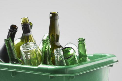 Several glass bottles of varying sizes and shades of green and clear, some with metallic or plastic caps, are placed inside a light green plastic bin, likely intended for bottle collection or recycling. The bottles are upright and appear to be empty, with some leaning against the sides of the bin, which is positioned on a flat surface against a plain white background. The bottles' surfaces exhibit different textures, with smooth, shiny glass reflecting light, and some showing visible ridges around their necks. The setting suggests an indoor or outdoor environment related to waste collection or recycling activities, relevant to rubbish removal services. The overall scene reflects a typical collection of used bottles ready for disposal or recycling, aligning with professional waste management practices by companies such as Commercial Waste Willesden.