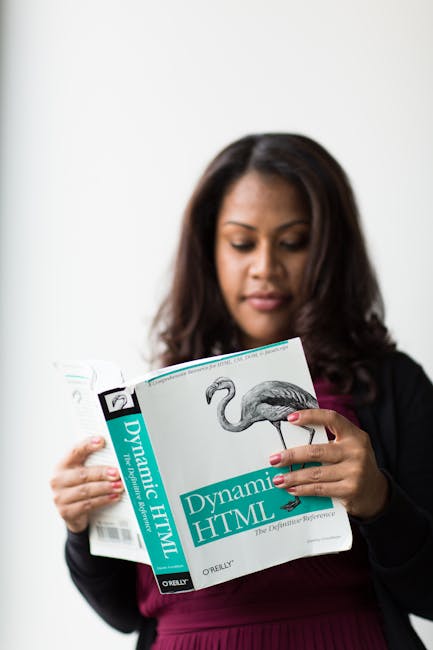 A woman with shoulder-length dark hair and medium skin tone is holding an open book titled 'Dynamic HTML' with a diagram of a flamingo on the cover. She is dressed in a dark blazer over a maroon top a