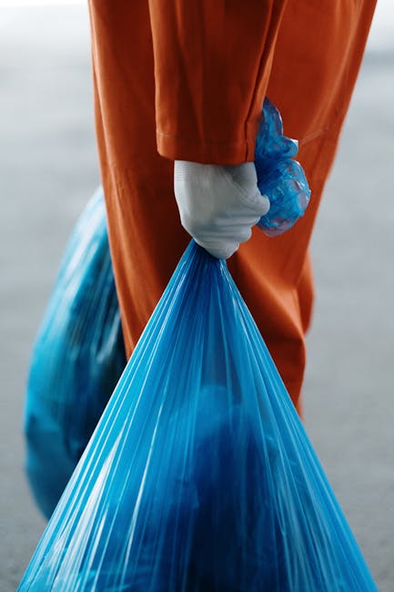 A person dressed in an orange protective suit and wearing white gloves is holding a large, blue, semi-transparent plastic bin bag filled with waste. The bag appears bulky and is tightly gathered at th