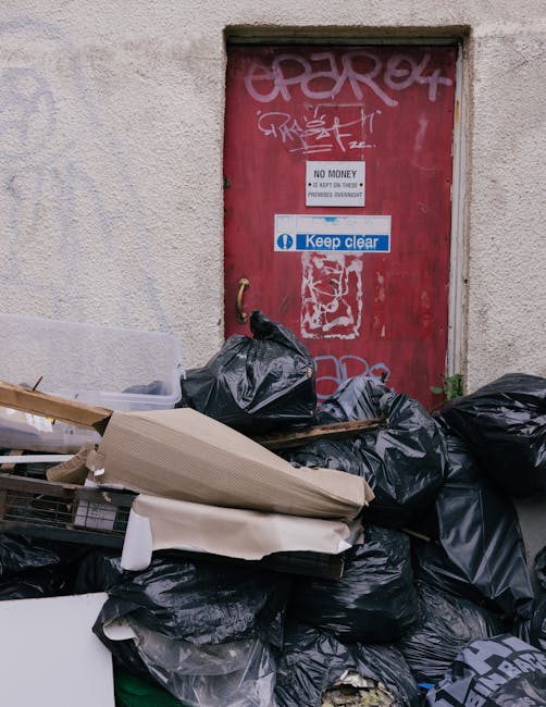 A person dressed in an orange protective suit and wearing white gloves is holding a large, blue, semi-transparent plastic bin bag filled with waste. The bag appears bulky and is tightly gathered at th