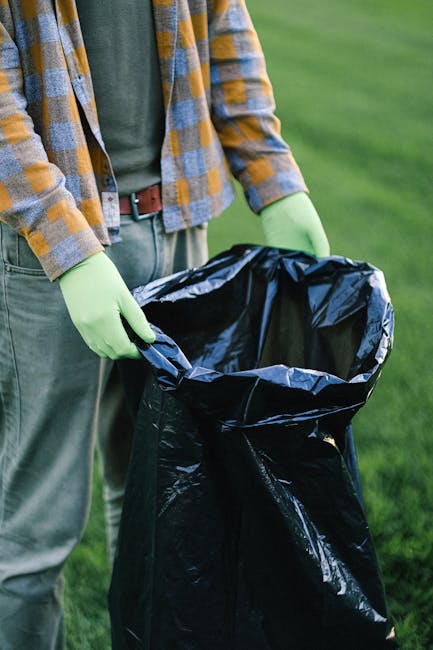 A person standing outdoors on a grassy area, dressed in a plaid shirt with shades of yellow and blue, over a green T-shirt, and wearing grey trousers with a brown belt. They are wearing light green gl