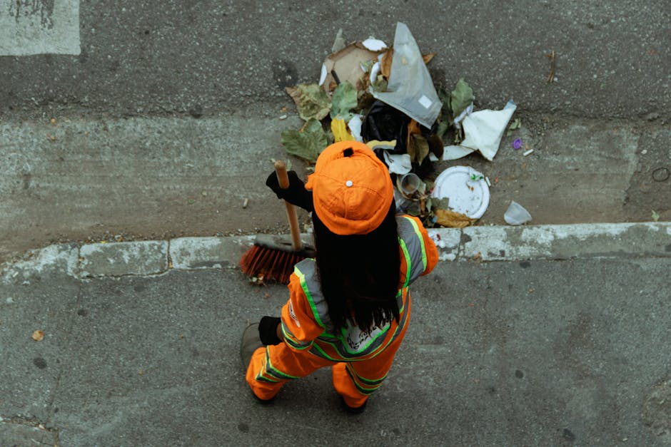 A pile of mixed domestic waste and rubbish consists of black plastic bin bags, cardboard boxes, an old, discarded beige car seat, and various small debris scattered on a gravel surface. The waste is p
